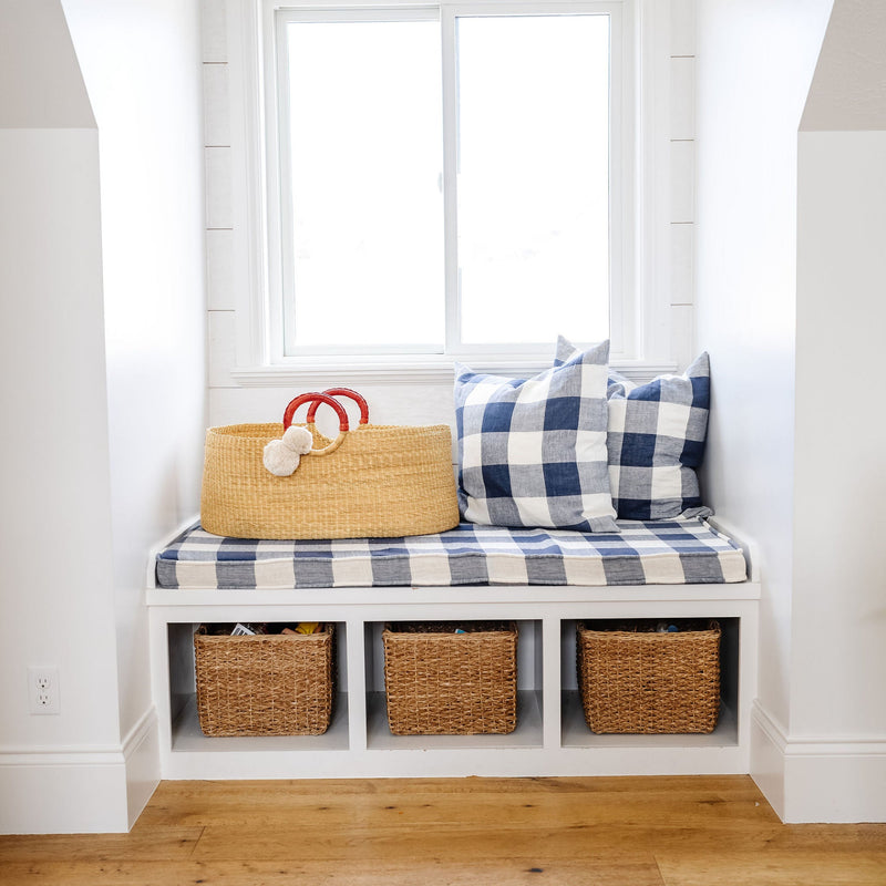Bench with plaid cushion, wicker basket, and decorative pillows in a room with wooden floor and white walls.